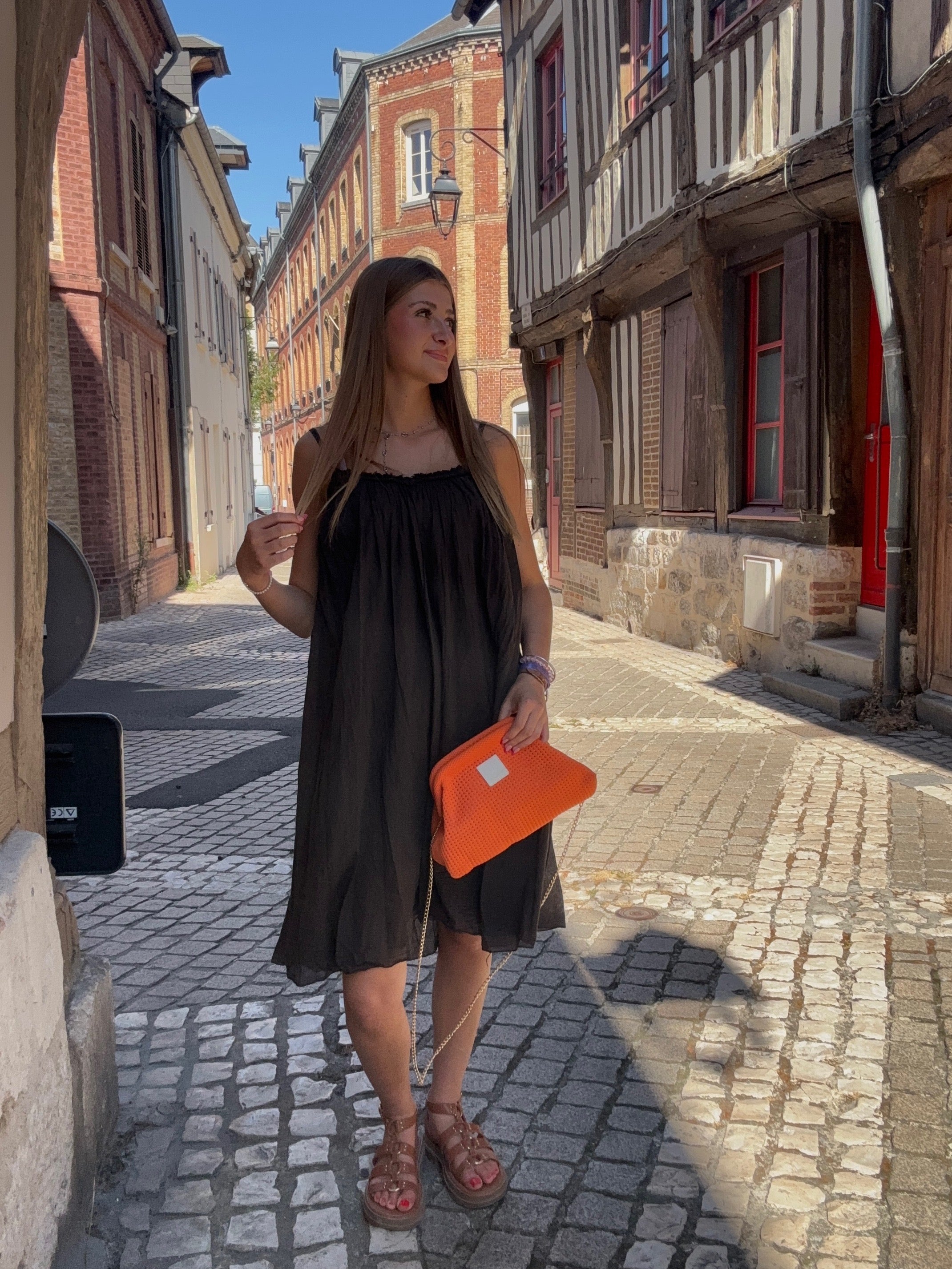 Robe mi-longue chocolat fluide à bretelles spaghetti portée avec un sac orange dans une ruelle pavée