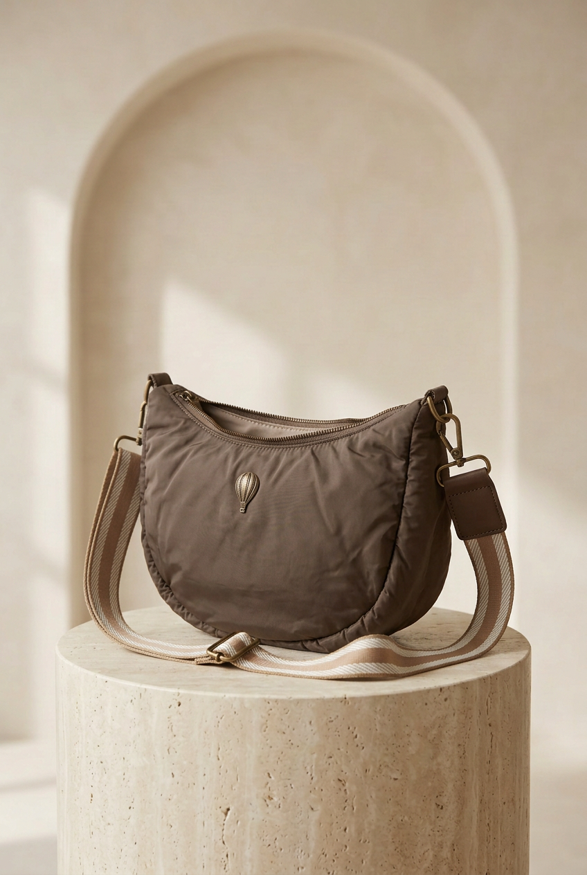 Sac demi-lune taupe avec bandoulière rayée, fermeture zippée et emblème montgolfière